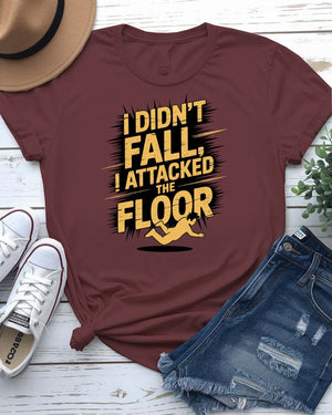 Funny unisex t-shirt that reads “I Didn’t Fall I Just Attacked The Floor” in bold meme font, modeled by man and woman in casual indoor setting.