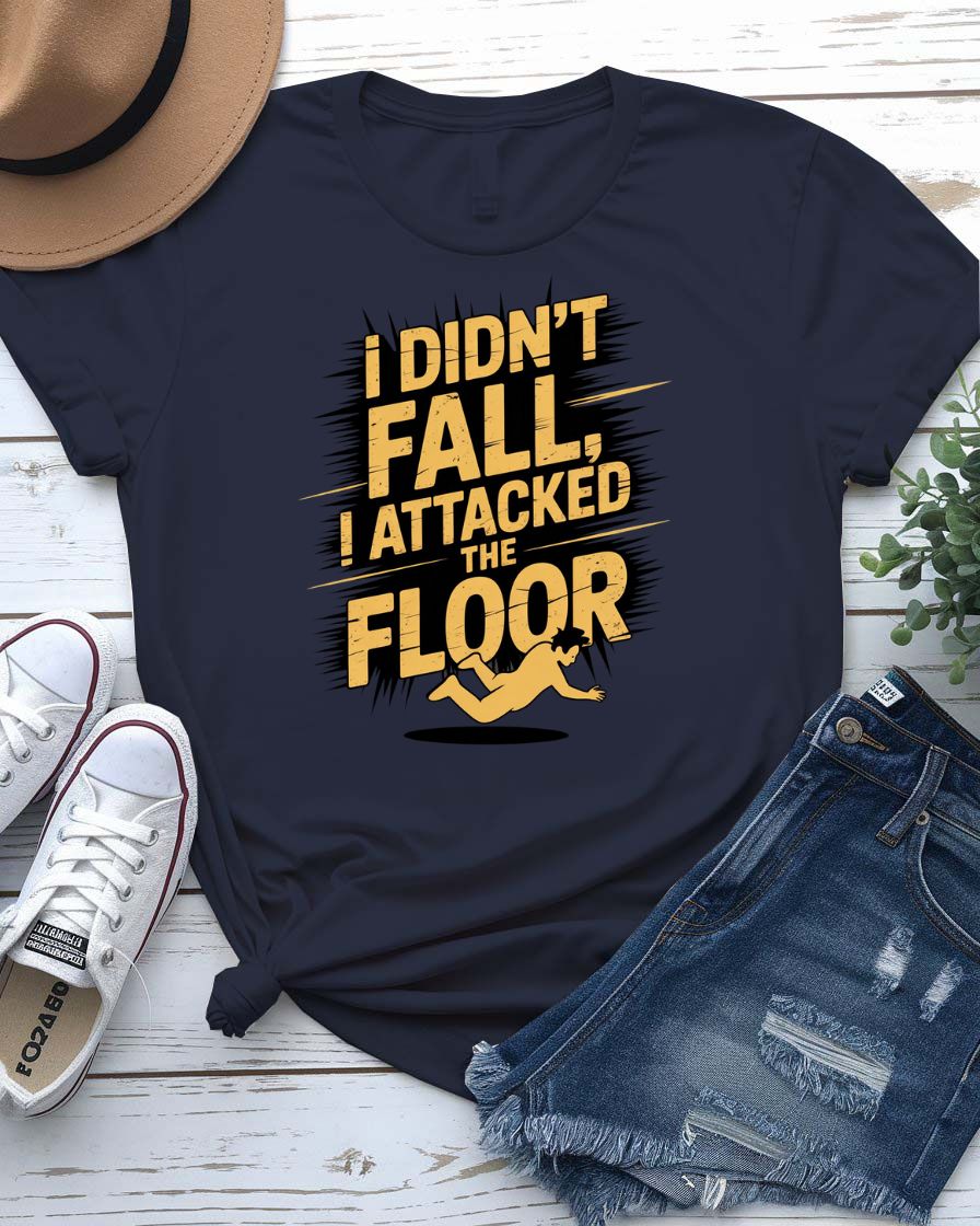Funny unisex t-shirt that reads “I Didn’t Fall I Just Attacked The Floor” in bold meme font, modeled by man and woman in casual indoor setting.