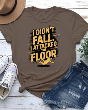 Funny unisex t-shirt that reads “I Didn’t Fall I Just Attacked The Floor” in bold meme font, modeled by man and woman in casual indoor setting.
