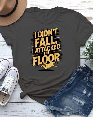 Funny unisex t-shirt that reads “I Didn’t Fall I Just Attacked The Floor” in bold meme font, modeled by man and woman in casual indoor setting.